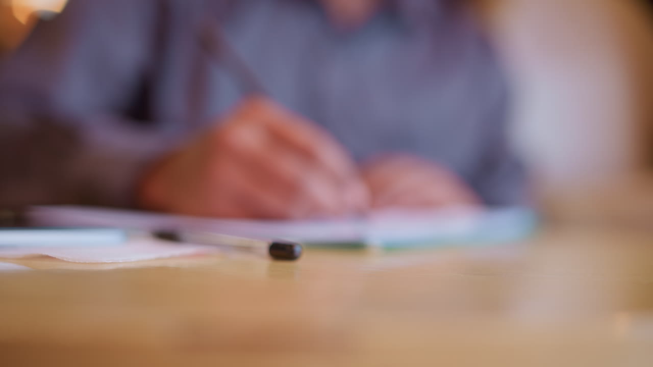 Blurred figure in background working at desk while pencil in foreground rests on wooden surface, hand raised mid-gesture suggests explanation or thought, creating mood of study, planning, or discussion