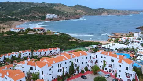 Vistas-Aéreas-Del-Tradicional-Hotel-De-Apartamentos-Blancos-Con-Techos-De-Tejas-Rojas,-Piscina-Y-Canchas-De-Tenis,-Con-Una-Hermosa-Cala-Al-Fondo-En-Menorca,-España