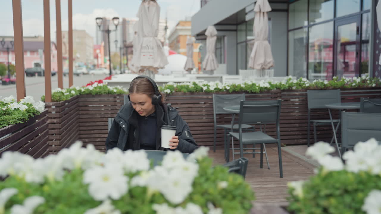 young woman walking onto cafe terrace, smiling and picking up coffee cup, relaxed posture and light movement, urban storefronts and floral planters framing scene, casual jacket and backpack, warm