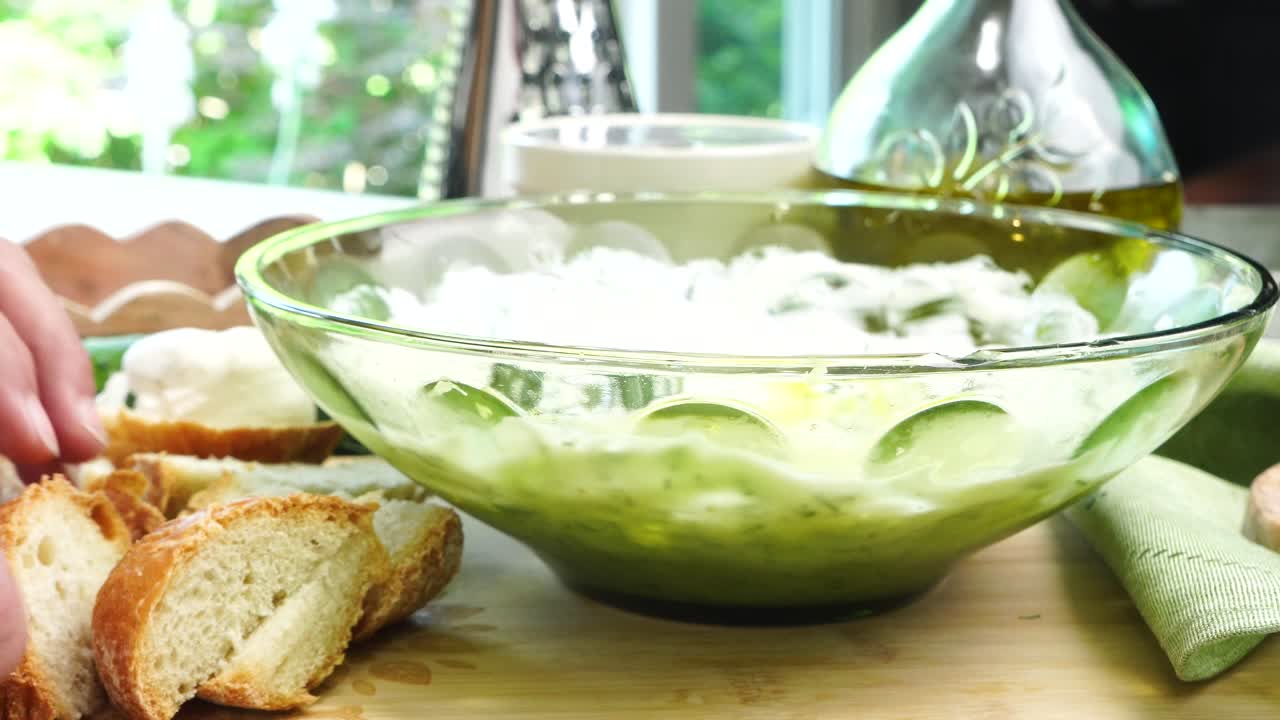 Placing bread on a wood cutting board. Making Tzatziki Greek or Mediterranean sauce or dip for a party. Dip or sauce in a green bowl on a wooden cutting board with dill, green napkin and a grater.