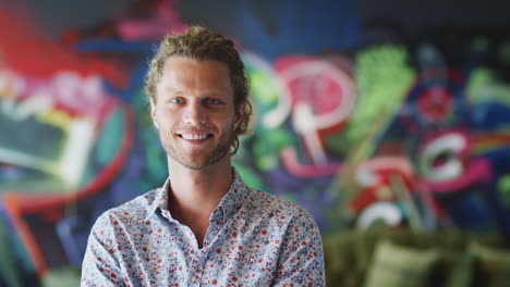 Young-white-male-creative-smiling-to-camera-in-front-of-mural-in-workplace,-head-and-shoulders