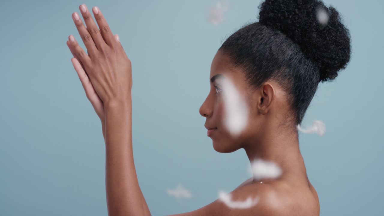 retrato de belleza en cámara lenta mujer afroamericana atractiva plumas cayendo sobre la piel lisa tocando hombros desnudos disfrutando del cuidado natural de la piel acariciando suavemente el cuerpo en fondo azul
