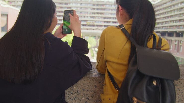Vista posteriore di due giovani amiche che visitano il Barbican Centre nella City di Londra e scattano una foto con il cellulare 3