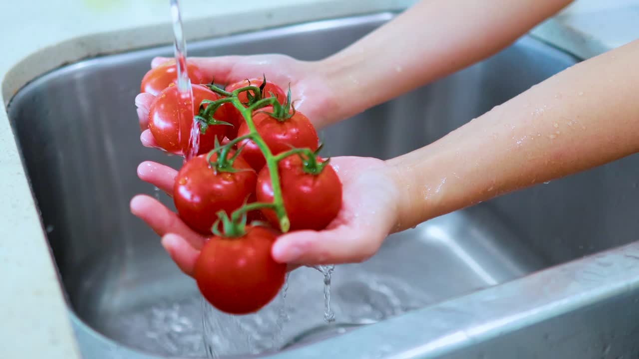 A person rinses vine tomatoes under a stream of water in a stainless steel kitchen sink, using natural lighting and steady close-up framing