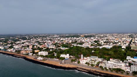 an expansive aerial shot captures the vibrant coastline and dense cityscape of pondicherry. the drone footage glides over the scenic beach, showcasing the famous promenade and an iconic monument
