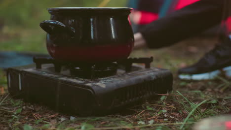 close up of cooking pot placed on portable gas stove with flame underneath set on forest floor during outdoor camping as grass and leaves surround stove in natural environment