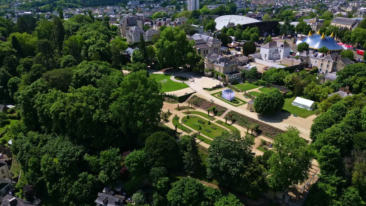 Lateral drone view of Jardin de la Perrine in Laval, with trees, circus tent, and urban backdrop in Mayenne, France.