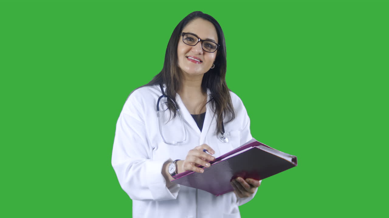 pantalla verde chromakey feliz mujer médico indio con gafas, bata de laboratorio y estetoscopio tomando notas en un clipboard