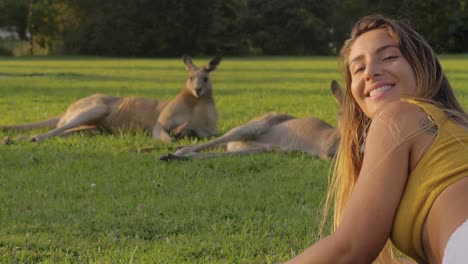 chica tendida en la hierba verde mirando a los canguros grises del este descansando en un día soleado - chica se da la vuelta y sonríe dulcemente a la cámara - costa dorada, qld, australia