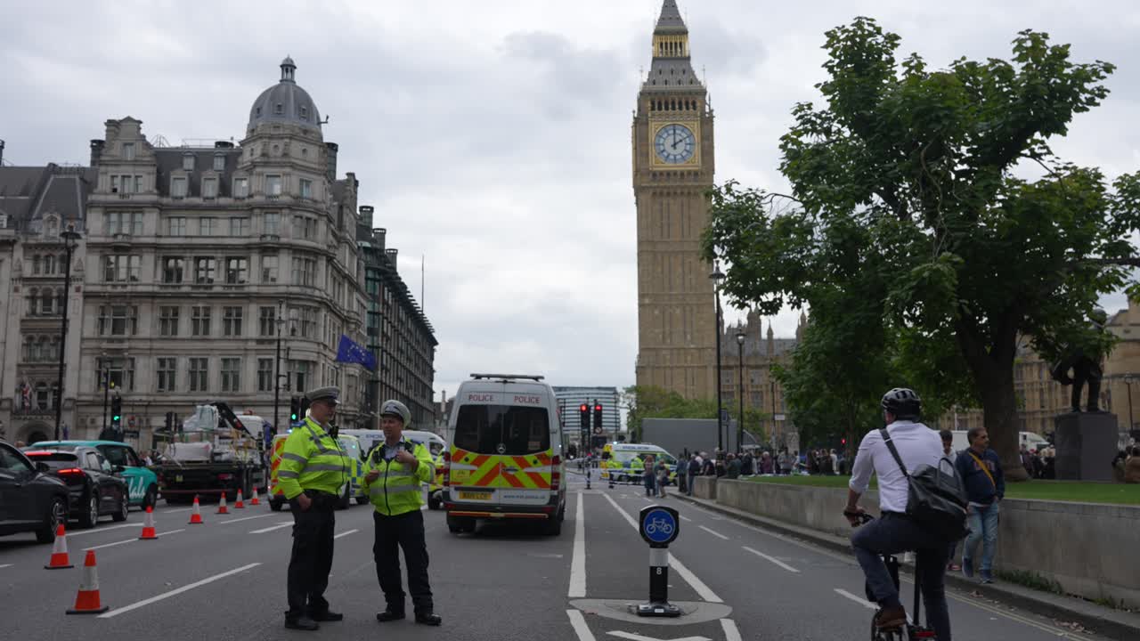 Two police officers stand in front of a van and red and white traffic cones marking the start of a cordon at the scene of a major incident in front of the Houses of Parliament.