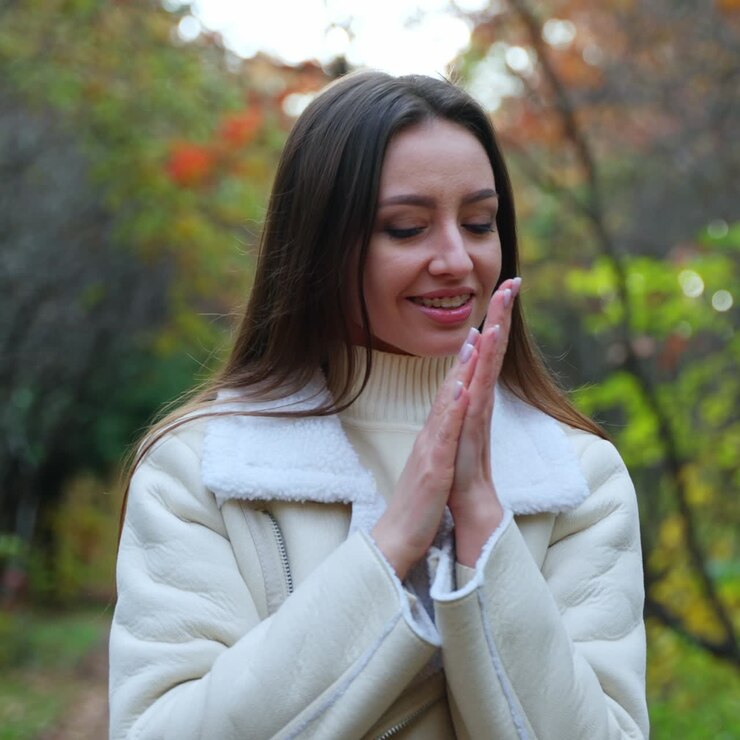 Beautiful rejoicing woman wearing white warm jacket taking a stroll by the forest. Lady warms her hands and enjoys fresh autumn air