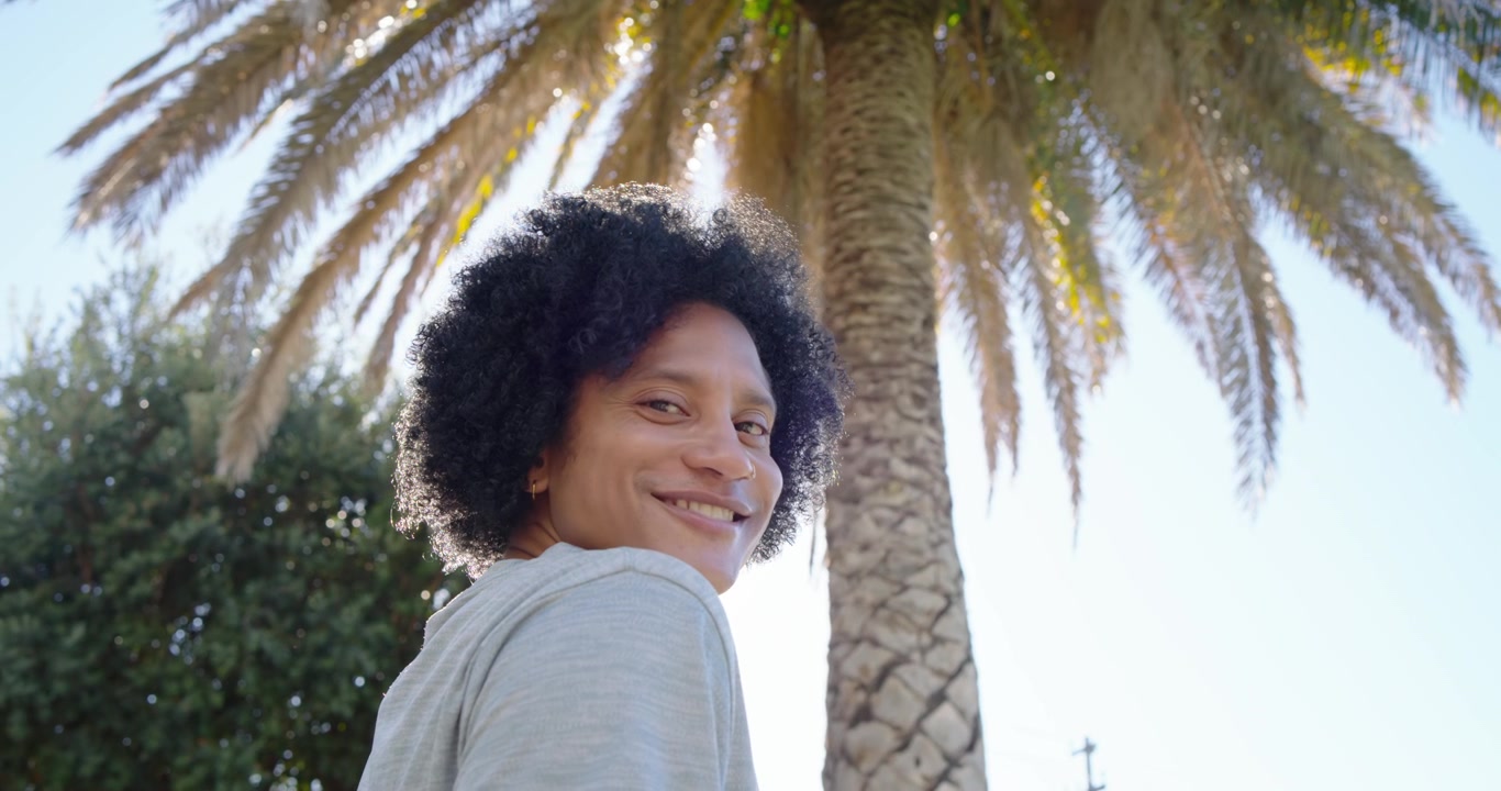 Homem afro-americano notando a câmera, passando por uma palmeira no parque, virando e sorrindo, com espaço para texto.