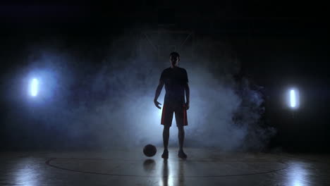 Basketball-player-in-sportswear-red-shorts-and-a-blue-t-shirt-goes-on-a-dark-basketball-court-in-the-backlighting-coming-out-of-the-smoke-knocks-a-basketball-ball-on-the-floor-looking-at-the-camera-in-slow-motion