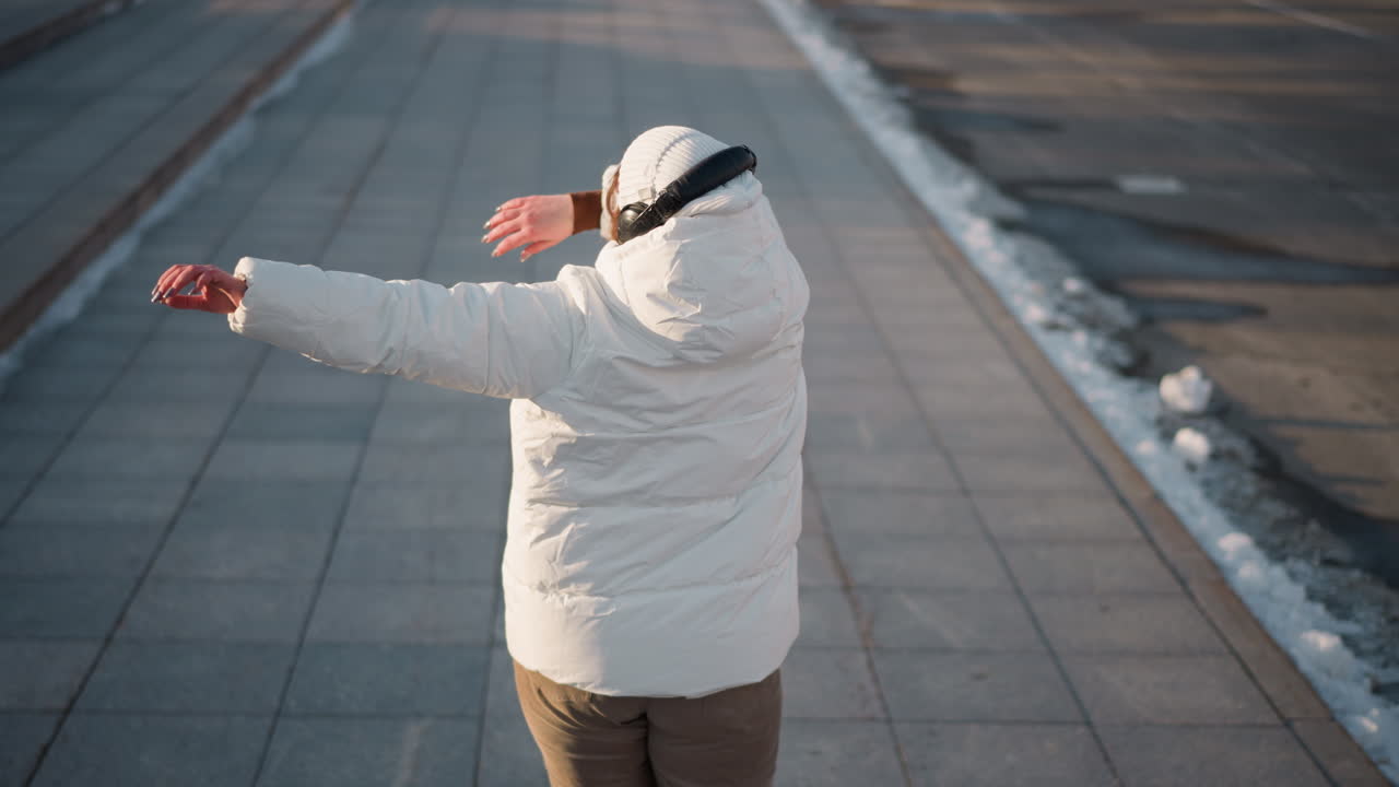 Back view of creative in white winter coat and beanie moving rhythmically on wide urban sidewalk during sunny winter day, enjoying peaceful moment with headphones under clear sky