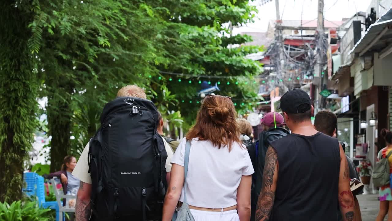 A group of friends walks down a leafy street lined with shops and greenery.