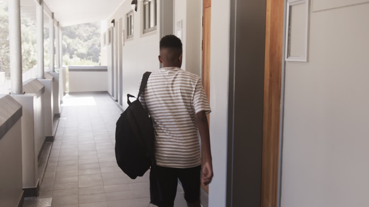 Walking down school hallway, African American teenage boy carrying backpack heading to classroom