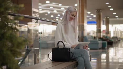 sophisticated lady seated on mall bench smiling and chatting on tablet in bright hall, casual jeans and sweater, black handbag at side, ambient ceiling lights and glass railings in background