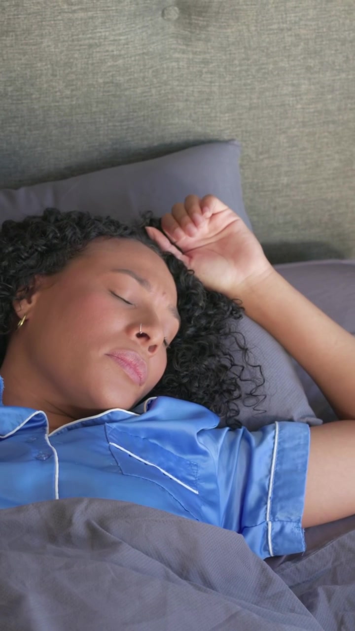 Vertical video: Yawning woman stretching, coming awake and smoothing blanket in bed in blue pajamas