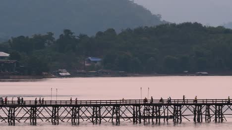 Mon-Bridge-and-a-longboat-followed-speeding-behind-the-structure-while-people-are-walking-on-the-bridge,-silhouetting-as-it-was-getting-dark,-in-slow-motion