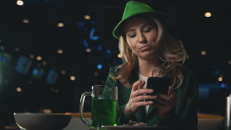 retrato de mujer rubia usando un teléfono inteligente, bebiendo una jarra de cerveza verde y usando un sombrero verde irlandés