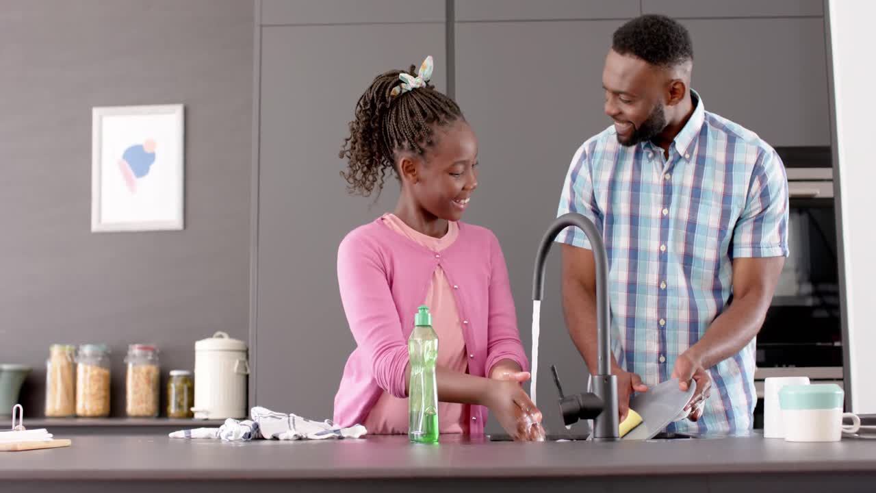 Happy african american father and daughter washing dishes and smiling in kitchen, slow motion