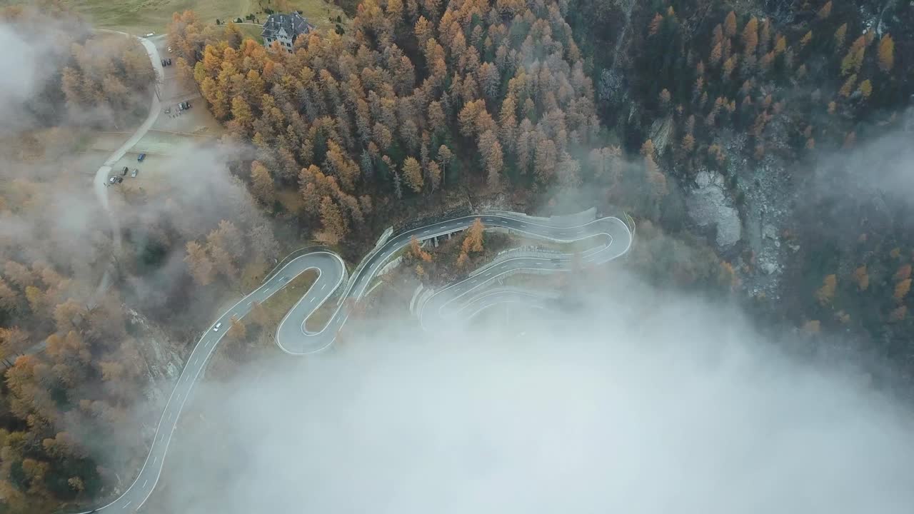 Aerial view of a winding road through a forested area, partially obscured by clouds, with autumn-colored trees surrounding the scene