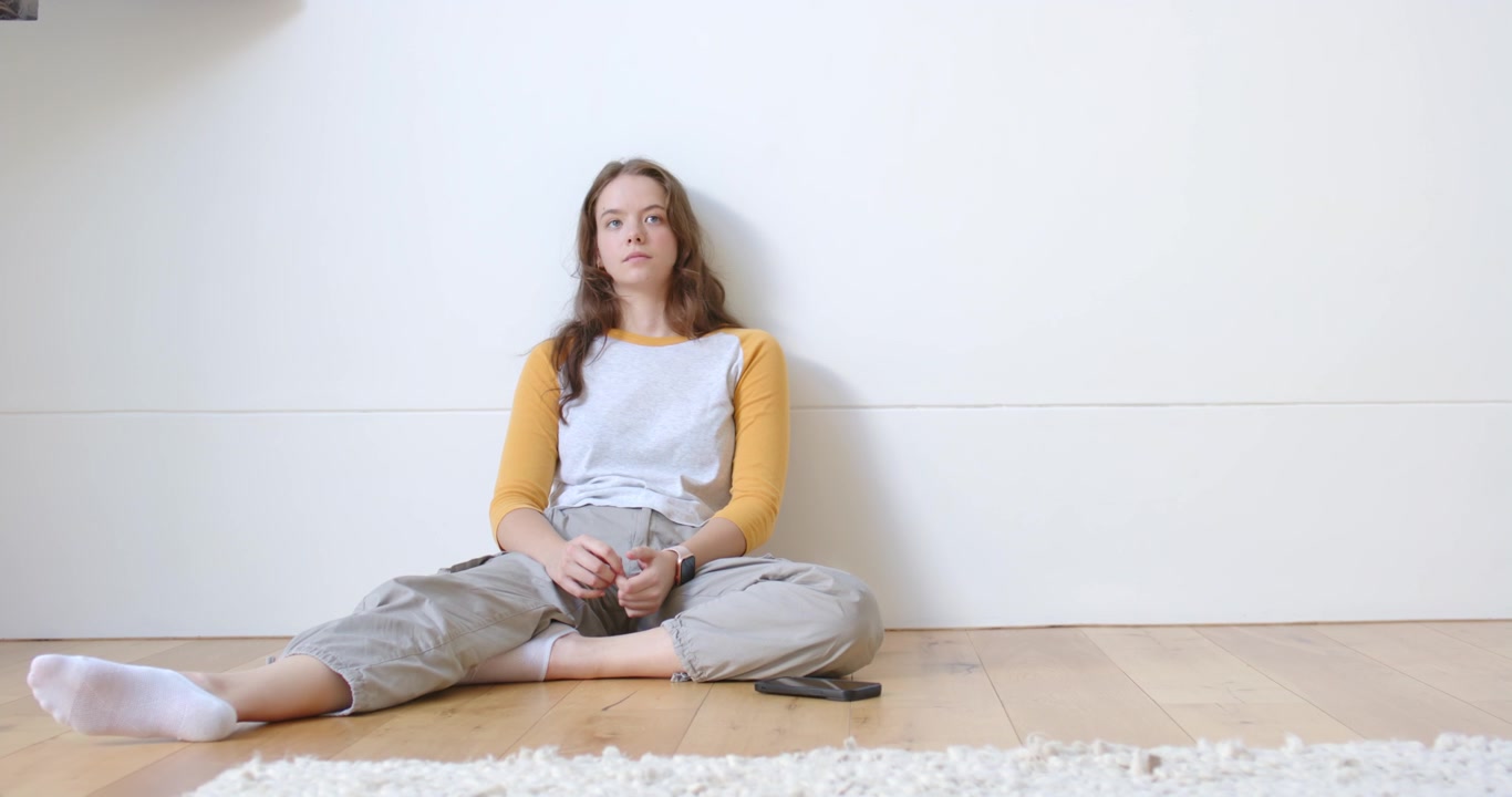 Woman sitting on wooden floor holding smartphone, shifting hands, glancing toward soft window light