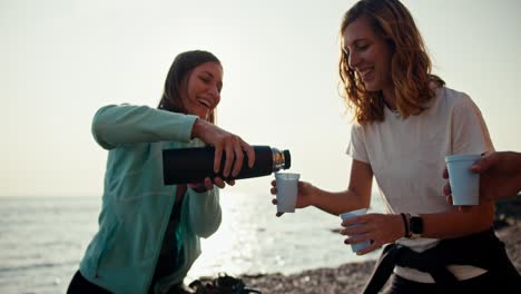 ein mädchen in einer blauen jacke gießt einem blonden mädchen mit einem weißen t-shirt während ihrer ruhe und entspannung während des klettern tee aus einer thermos. eine gruppe von klettern trinkt tee während ihrer pause zwischen den klettern