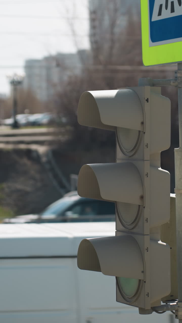 Close view of a city traffic light and pedestrian crossing sign in an urban area, with cars passing by and someone standing by the road pavement