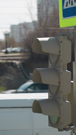 vista cercana de un semáforo de tráfico de la ciudad y un letrero de cruce de peatones en un área urbana, con coches pasando y alguien de pie junto al pavimento de la carretera