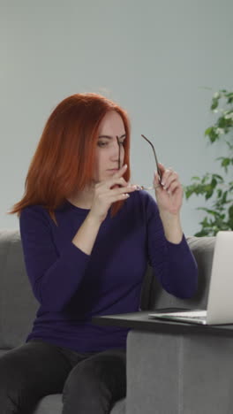 freelancer with red hair sits on sofa putting on glasses to continue work on laptop. young woman uses modern electronic device in living room slow motion