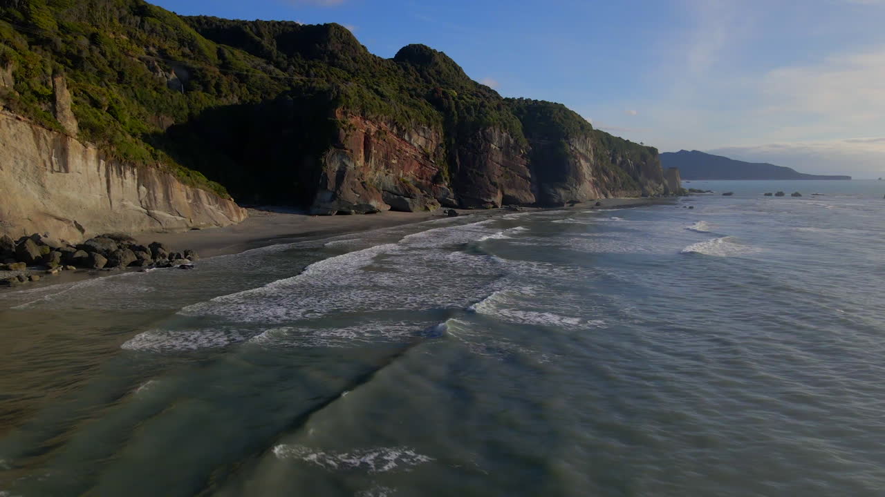 Waves splashing ashore a wild sandy beach with steep cliffs by Tasman sea in New Zealand