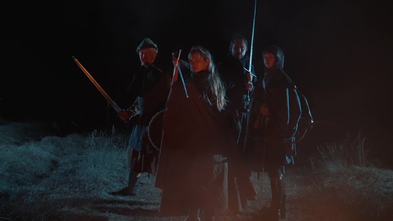 Fyra bepansrade krigare rör sig mot kameran genom mörk dimma, med den centrala, mantlade ledaren som lyfter svärdet upprätt medan följeslagarna håller spjut och runda sköldar, vilket skapar dramatisk stridsspänning.