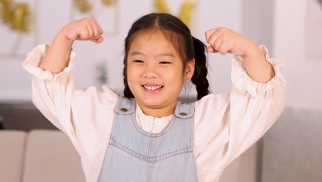 young asian girl with pigtails confidently flexes her arms and smiles in a well-lit, modern living room. soft, natural lighting enhances cheerful mood