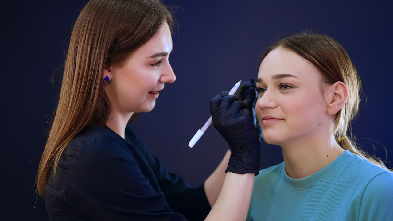 Smiling beauty consultant turns the girl's head in a necessary position. Cosmetologist is going to mark the brow line with white pen.
