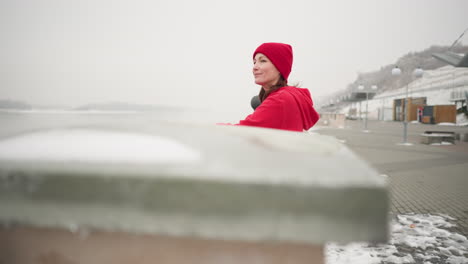 woman in red hoodie rests on iron fence with hands on decorative metal railing, gazing thoughtfully at calm water environment, misty atmosphere with snow-covered ground and distant shoreline