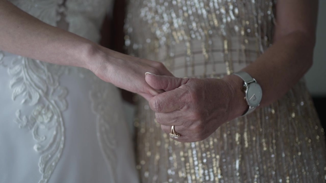 Primer plano de dos mujeres tomadas de la mano, una con un vestido de novia y la otra con un vestido de lentejuelas, mostrando un momento de conexión.