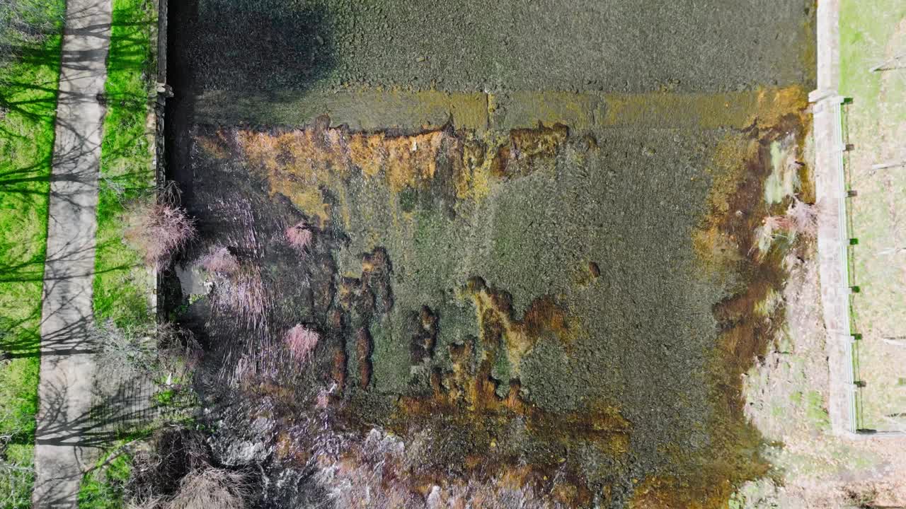Drohnenaufnahme von oben eines flachen Flusses mit sichtbaren Flussbettsteinen, Algen und fließendem Wasser zwischen Uferböschungen in Spanien. Natürliche Süßwassertextur und Landschaftshintergrund.