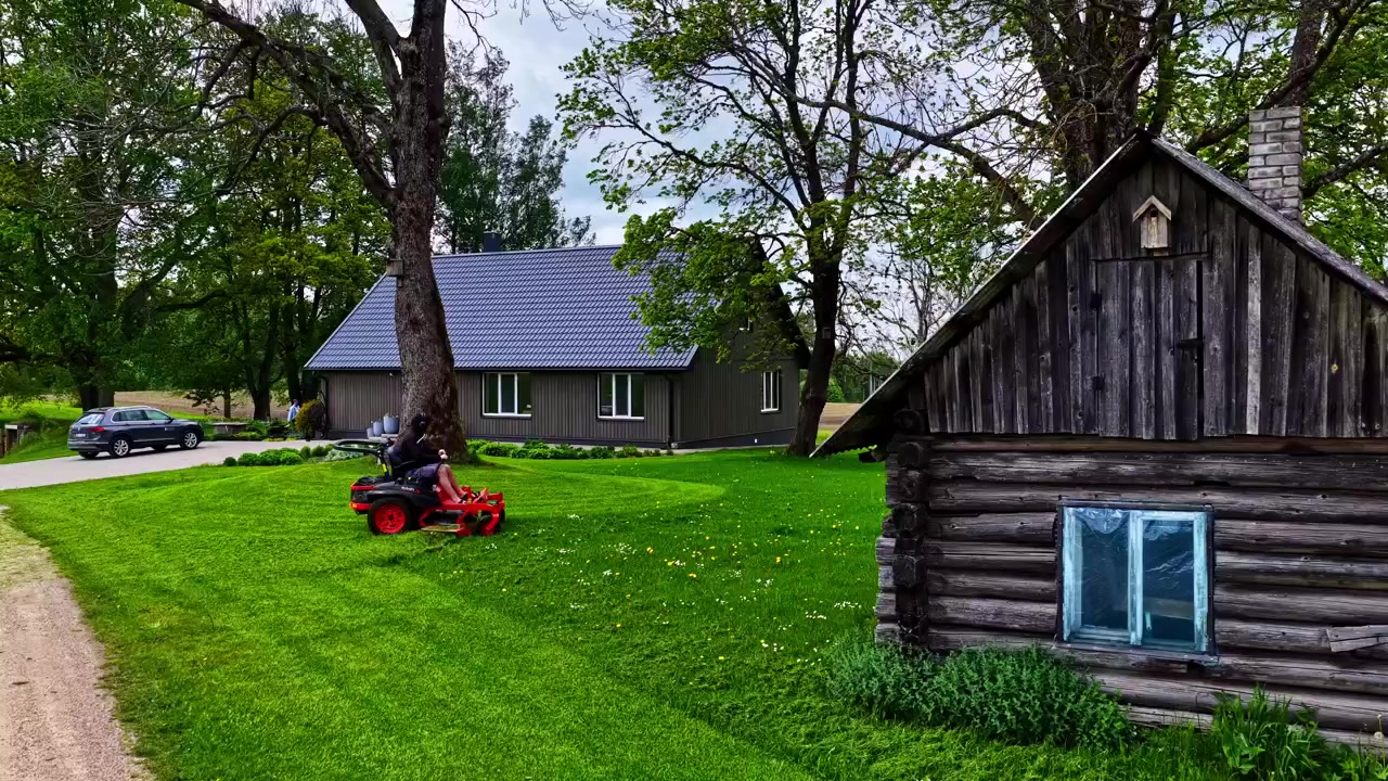 Person på rød havetraktor klipper frodig grøn græsplæne mellem moderne gråt hus og gammel rustik trælade med store træer i landlige Jelgava Letland på en sommerdag