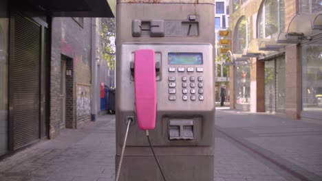 Tiro-De-Muñeca-De-Un-Viejo-Teléfono-Público-De-Color-Rosa-Con-Manchas-Oxidadas-En-La-Calle-Comercial-De-Colonia,-Alemania,-En-Un-Hermoso-Día-Soleado