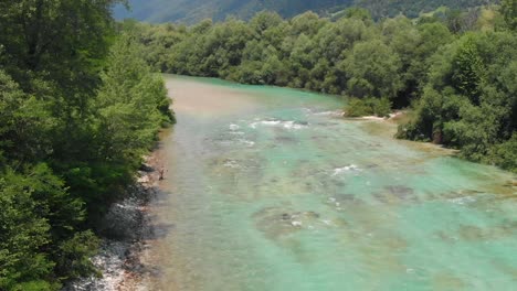 person fishing in fresh flowing turquoise river alongside lush forest woodland rising aerial view