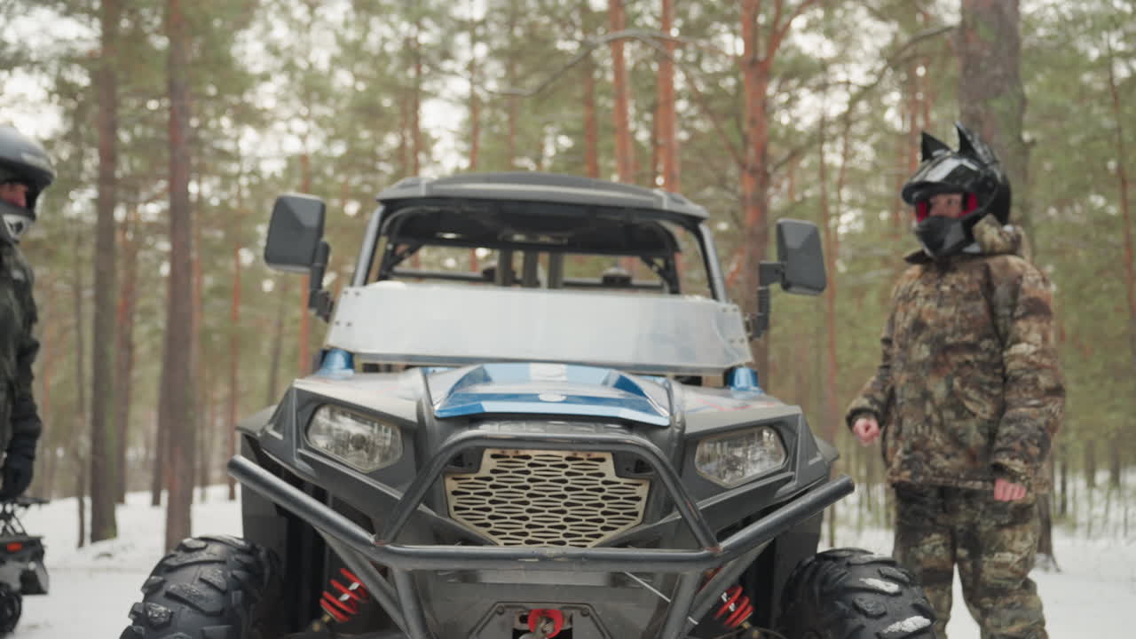 Ispezione ravvicinata della griglia anteriore del quad, i piloti controllano paraurti e luci, due figure con casco esaminano le sospensioni, cassetta degli attrezzi e verricello sulla neve, intesa meccanica con stretta di mano della squadra, inquadrature dettagliate del veicolo.