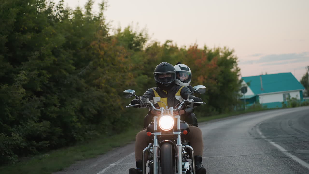 Couple wearing helmets rides motorcycle on countryside road during evening sunset, passing trees and houses, enjoying freedom, adventure, and peaceful atmosphere of rural landscape