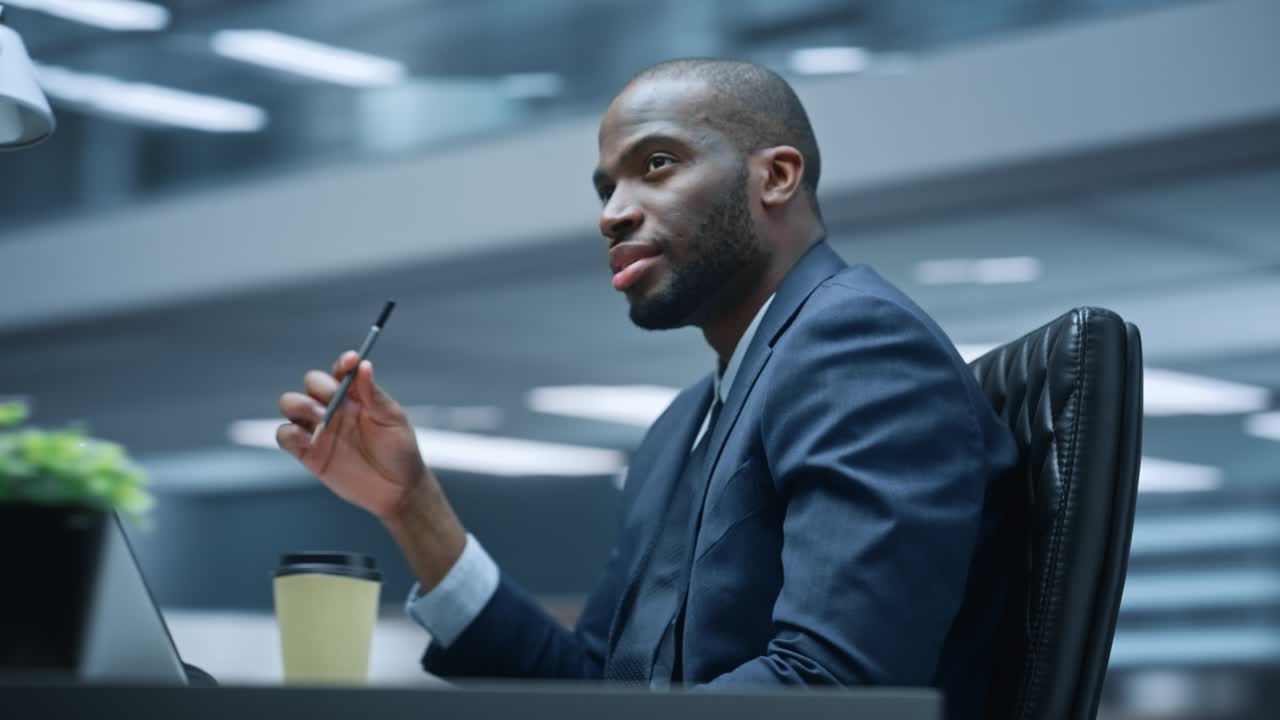 360 graders kontor: Succesfuld sort forretningsmand sidder ved skrivebord og tænker, bruger bærbar computer. Afroamerikansk iværksætter udfører aktiemarkedsanalyse. Bevægelig sporingsoptagelse.