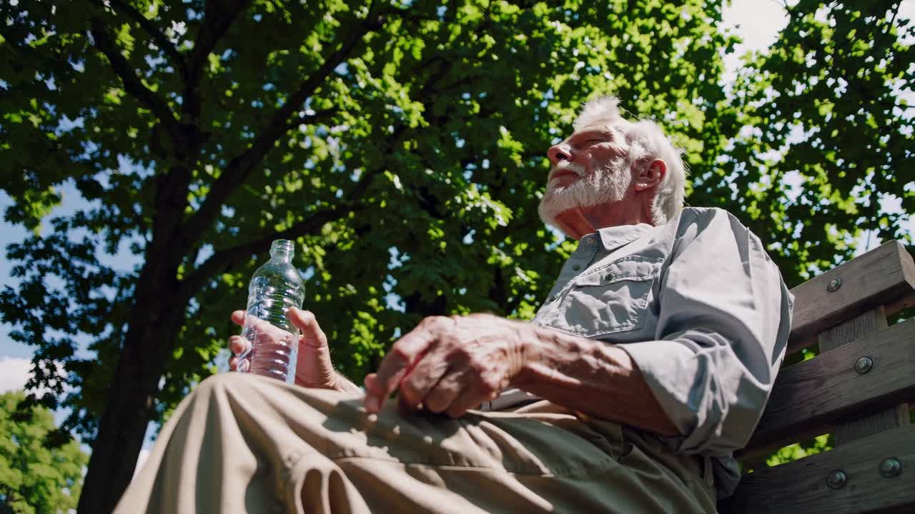 Low-angle video shot of a man sitting on a bench, drinking water under sunlit trees