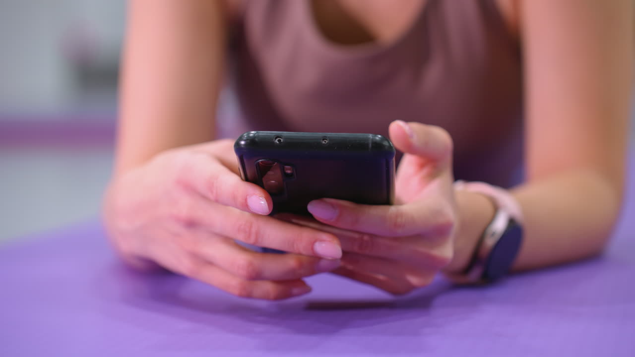primer plano de las manos sosteniendo un teléfono inteligente mientras se acuesta en una alfombra de yoga púrpura, la persona usa un reloj inteligente y tiene las uñas cuidadosamente cuidadas, la iluminación suave destaca la tecnología moderna