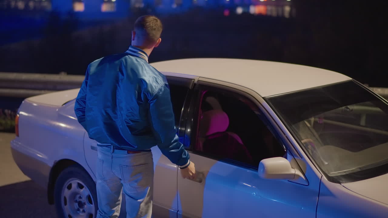 Ung, kaukasisk mand nærmer sig parkeret sedan i vejsiden, inspicerer dør og interiør under neonlys, forsigtig stilling i blå jakke, filmisk spænding og urban noir-atmosfære