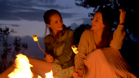 two young caucasian girls sitting by fire in evening in nature, preparing a marshmelow, looking at open fire, thinking