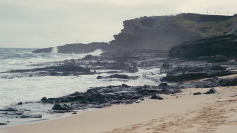 Sandy-beach-landscape-with-waves-splashing-on-rocks-and-cliffside-in-the-background