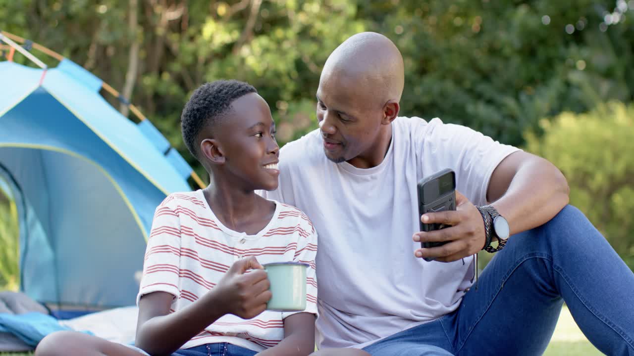 African father and youth son tilting smartphone, son holding mug, smiling on blanket by blue tent
